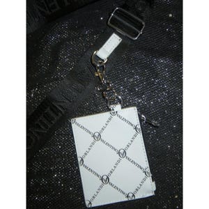 May include: White leather cardholder with the "Valentino Orlandi" logo in a repeating pattern. The cardholder is attached to a silver-tone metal clasp and a white leather strap. The background is a black, textured surface.