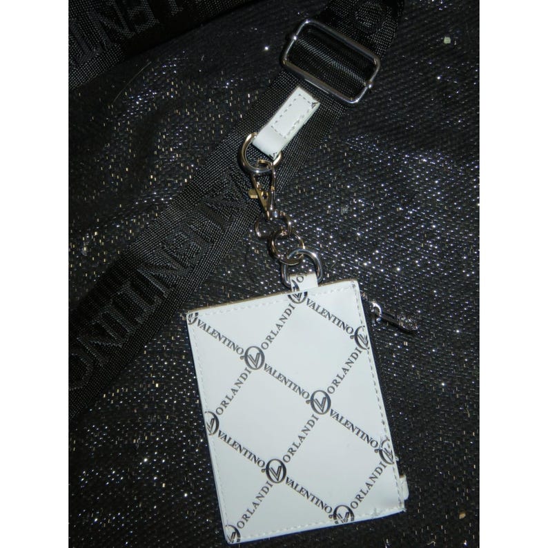 May include: White leather cardholder with the "Valentino Orlandi" logo in a repeating pattern. The cardholder is attached to a silver-tone metal clasp and a white leather strap. The background is a black, textured surface.