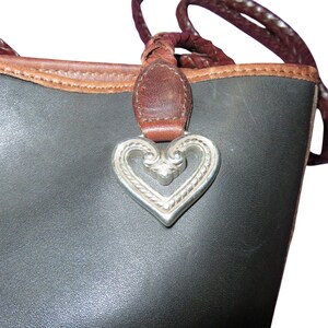 May include: Close-up of a black leather tote bag with a brown leather trim and braided handles. A silver heart-shaped charm hangs from the bag's handle, adding a decorative touch to the design.