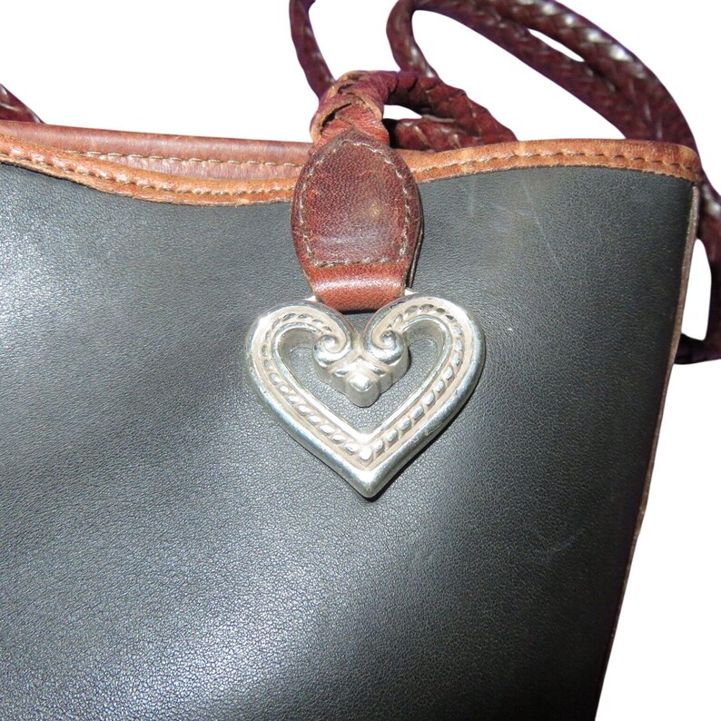 May include: Close-up of a black leather tote bag with a brown leather trim and braided handles. A silver heart-shaped charm hangs from the bag's handle, adding a decorative touch to the design.