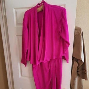 Puede incluir: Un conjunto de dos piezas fucsia brillante, que consta de una chaqueta de manga larga y una falda a juego. La chaqueta tiene un frente drapeado y la falda parece ser hasta la rodilla. El conjunto está colgado en una puerta blanca, con un abrigo beige a la derecha.