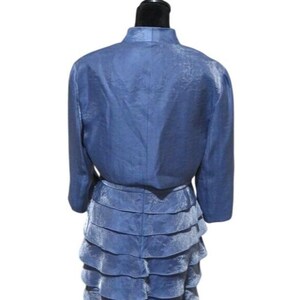 May include: A two-piece blue dress set. The jacket has long sleeves and a mandarin collar. The skirt is tiered with multiple layers. The fabric has a subtle sheen. The dress is displayed on a black mannequin.