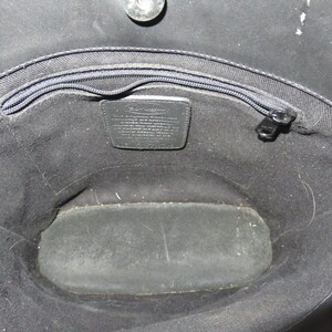 May include: Open view of a black handbag interior. The bag features a zippered top, a small leather tag with text, and a rectangular base. The interior is lined with black fabric.