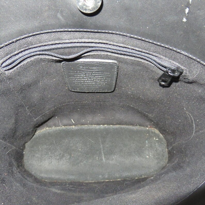 May include: Open view of a black handbag interior. The bag features a zippered top, a small leather tag with text, and a rectangular base. The interior is lined with black fabric.