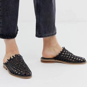 May include: Black woven leather slip-on mules with a low heel. The open-back design features a textured, open weave pattern and a light brown sole. The shoes are shown on a person wearing black denim jeans.