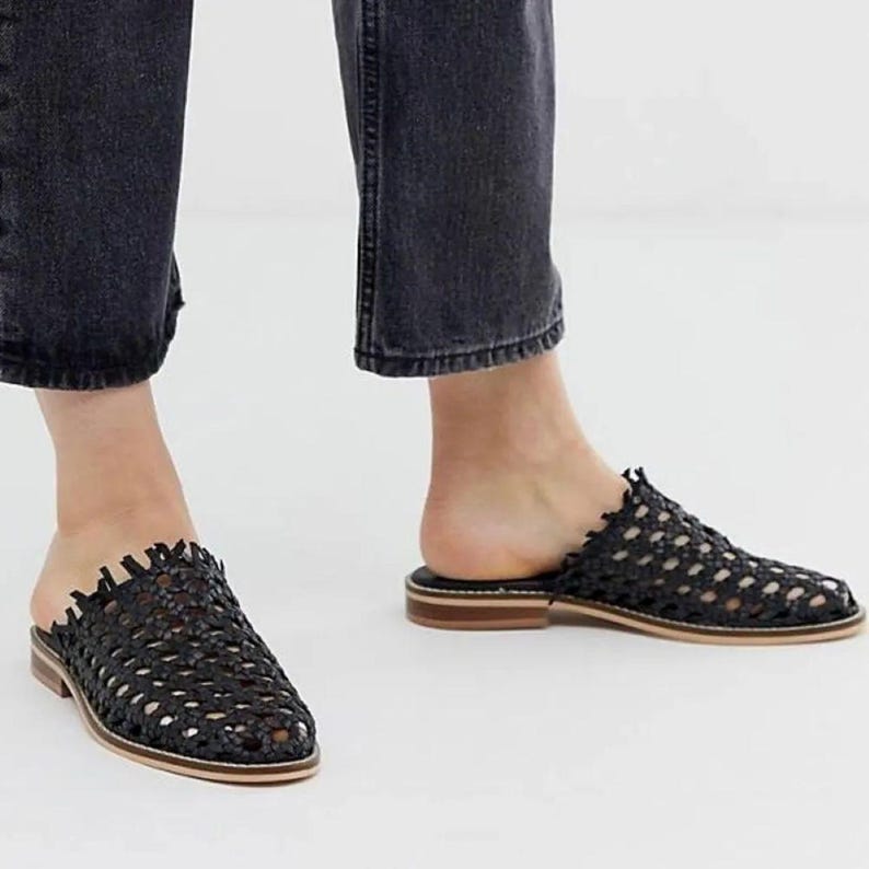 May include: Black woven leather slip-on mules with a low heel. The open-back design features a textured, open weave pattern and a light brown sole. The shoes are shown on a person wearing black denim jeans.