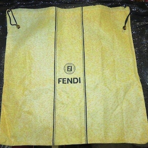 May include: A yellow drawstring bag with black vertical stripes and the Fendi logo. The bag has a textured surface and black drawstrings with gold-colored tips.