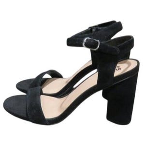 May include: Black suede sandals with a block heel. The sandals feature an ankle strap with a silver buckle and a strap across the toes. The heel height is approximately 3 inches.