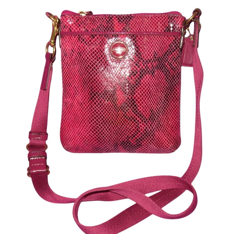 Peut inclure: Un sac bandouli&egrave;re rose fuchsia avec un motif peau de serpent. Le sac a une longue bandouli&egrave;re r&eacute;glable et une petite poche avant. Des d&eacute;tails dor&eacute;s ornent le sac, y compris la fermeture &eacute;clair et les attaches de la bandouli&egrave;re. Un logo circulaire est centr&eacute; sur le devant.