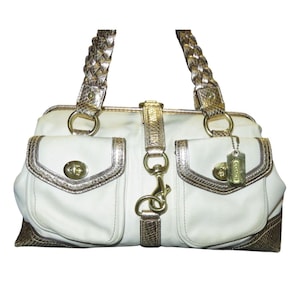 May include: Cream-coloured handbag with gold-tone hardware and accents. Features two front pockets with turn-lock closures, braided handles, and a detachable tag. The bag has a structured shape and a snakeskin-like trim.