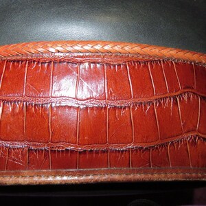 May include: Close-up of a brown leather belt with a crocodile skin texture. The belt has a braided leather trim along the top edge and a stitched border at the bottom. The leather is a rich, reddish-brown color.