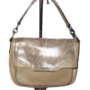 Peut inclure: Un sac à main beige avec un rabat à motif peau de serpent et une barre métallique argentée. Le sac a une bandoulière et un fond incurvé. Le sac est présenté sur un support métallique.