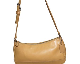 May include: A tan leather shoulder bag with a long adjustable strap. The bag has a zip closure and a small tag attached to the side. The bottom of the bag is a slightly darker shade of tan.