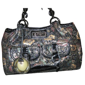 May include: A handbag featuring a dark, multi-colored metallic textured pattern with black patent leather trim. It includes two front pockets with buckle closures, a rectangular brand plaque, and a circular charm with a cream-colored center.