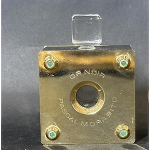 May include: A gold square perfume bottle with a clear glass stopper. The bottle has a green glass accent on each corner and the text "OR NOIR PASCAL MORABITO" on the front.