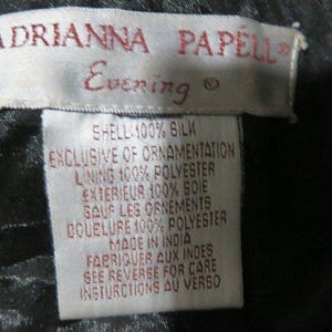 Op de afbeelding: Close-up van een wit label met rode tekst van Adrianna Papell Evening. Het label geeft de stof samenstelling: buitenkant 100% zijde, voering 100% polyester. Gemaakt in India. Zie achterkant voor onderhoudsinstructies.