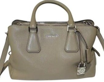 May include: A taupe-colored handbag with a top handle and a detachable shoulder strap. The bag features a silver zipper, silver hardware, and a small lock with a logo. The brand name "MICHAEL KORS" is visible on the front of the bag.