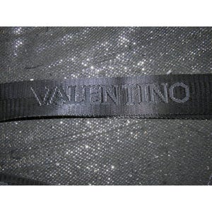 May include: Close-up of a black woven strap with the name "VALENTINO" embossed across it. The strap is set against a sparkly, textured background, creating a contrast in textures and tones. The overall aesthetic is sleek and modern.