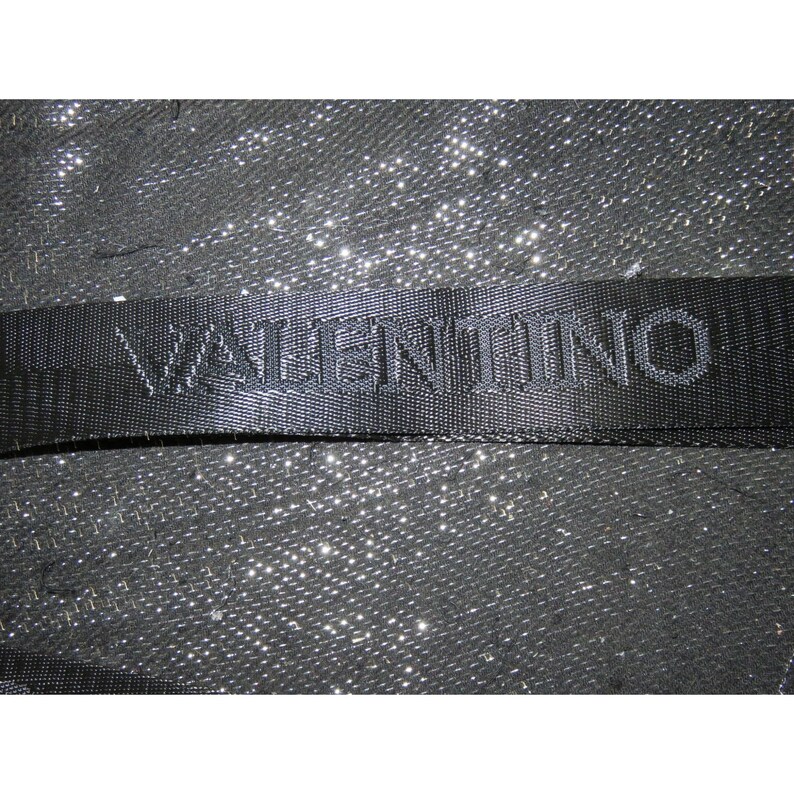 May include: Close-up of a black woven strap with the name "VALENTINO" embossed across it. The strap is set against a sparkly, textured background, creating a contrast in textures and tones. The overall aesthetic is sleek and modern.