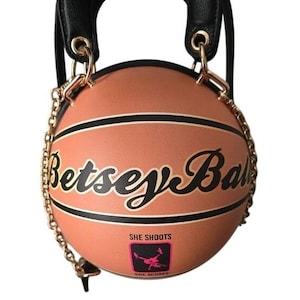 May include: A basketball-shaped handbag with a black handle and gold chain strap. The bag is orange with black stripes and the words "Betsey Ball" in cursive. A pink square with a silhouette of a person shooting a basketball is below the text.