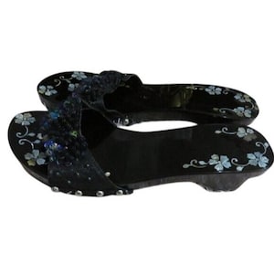 May include: Black wooden clogs with floral accents. The sandals feature a dark base with light blue and white flower designs. The straps are embellished with sequins and beads. The clogs have a slight heel and are decorated with silver studs.