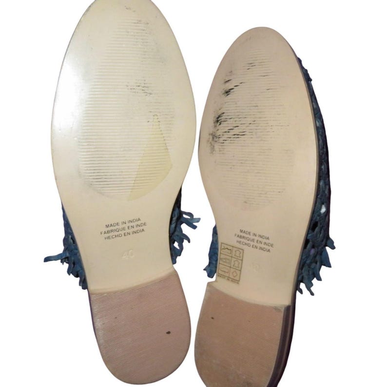 May include: A pair of blue woven flats with fringe detailing. The soles are light beige with the text "MADE IN INDIA FABRIQUE EN INDE HECHO EN INDIA" and size 40. The shoes have a low heel.