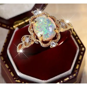 May include: A gold-toned ring with an oval opal gemstone at the center. The opal displays a play of color with iridescent hues. The ring is set with small, clear gemstones and intricate filigree detailing. The ring is presented in a red jewelry box.