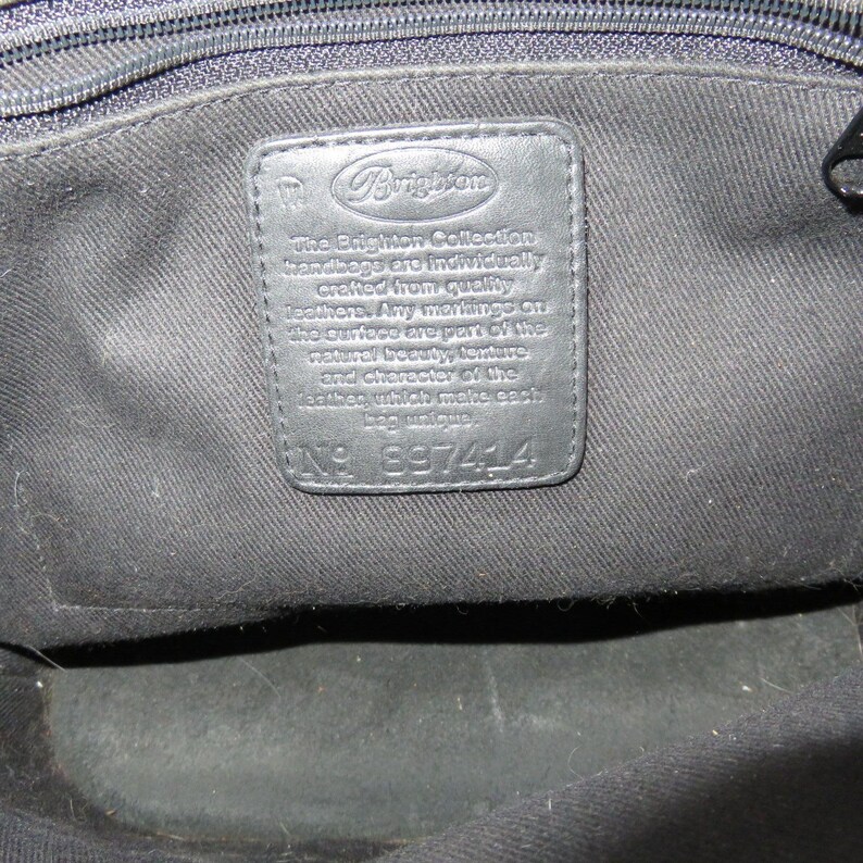 May include: Black leather handbag interior with a stitched leather tag. The tag features the Brighton logo and text that reads "The Brighton Collection handbags are individually crafted from quality leathers." The tag also has the number 897414.