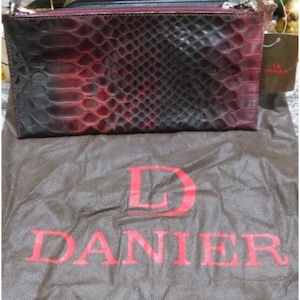Pode incluir: Uma clutch retangular com um padrão de pele de cobra em tons de vermelho e preto. A clutch tem um fecho de correr e uma etiqueta que diz "DANIEL". A clutch está em cima de uma bolsa castanha com o nome "DANIEL" impresso a vermelho.