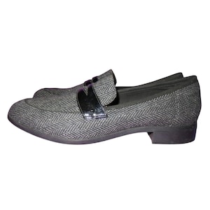 May include: A pair of gray herringbone pattern loafers with a black patent leather strap across the top. The shoes have a low heel and a rounded toe. The shoes are made of a woven fabric.