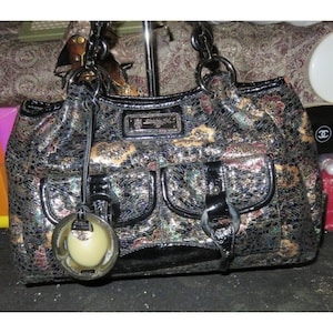 Op de afbeelding: Een zwarte handtas met een gouden en zilveren patroon. De tas heeft een grote, ronde, crèmekleurige hanger eraan. De tas heeft een zwarte leren band en een zwarte gesp.