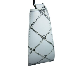 May include: White handbag with a repeating black pattern of the words "VALENTINO ORLANDI" and a logo. The bag has a silver-colored metal handle and a white strap. The bag is made of a smooth, shiny material.