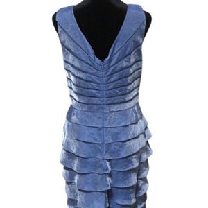 May include: A sleeveless, knee-length dress in a dusty blue hue. The dress features a V-neck and a tiered, ruffled skirt. The fabric has a subtle sheen, and the dress is displayed on a black mannequin.