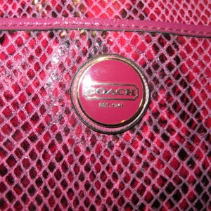 Peut inclure: Gros plan sur un sac &agrave; main Coach rose avec un motif peau de serpent. Le sac comporte un embl&egrave;me circulaire avec le logo Coach et le texte "EST. 1941". Le sac a une surface brillante et textur&eacute;e.