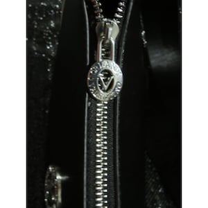May include: Close-up of a black handbag with a silver zipper. The zipper pull has a circular design with the letter "V" in the center. The bag appears to be made of a shiny material.