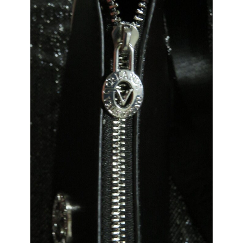 May include: Close-up of a black handbag with a silver zipper. The zipper pull has a circular design with the letter "V" in the center. The bag appears to be made of a shiny material.