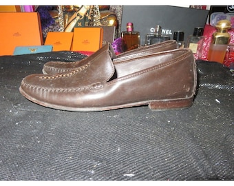 Puede incluir: Un par de mocasines de cuero marrón con un detalle cosido alrededor de la puntera. Los mocasines tienen un diseño de deslizamiento y una puntera ligeramente puntiaguda.