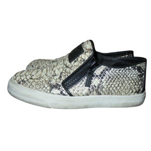 May include: A pair of slip-on sneakers with a snakeskin pattern in shades of beige, brown, and black. The sneakers feature a black zipper detail on the side and a white rubber sole.