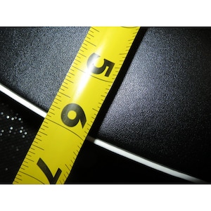 May include: A yellow tape measure with black markings, showing the numbers 5, 6, and 7 in inches. The tape measure is angled across a textured black surface, with a white edge visible.
