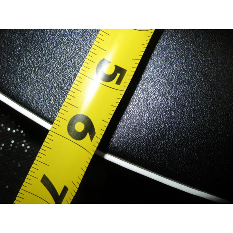 May include: A yellow tape measure with black markings, showing the numbers 5, 6, and 7 in inches. The tape measure is angled across a textured black surface, with a white edge visible.