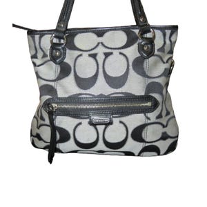 May include: Coach tote bag in a gray and black pattern. The bag features a zip closure, black leather handles, and a front zip pocket. The Coach logo is visible on the front of the bag.