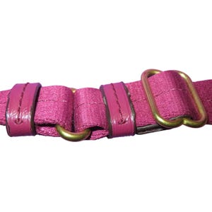 Peut inclure: Gros plan sur une sangle magenta avec une garniture en m&eacute;tal dor&eacute;. La sangle est en tissu tiss&eacute; avec des accents de cuir magenta brillant. La quincaillerie comprend une boucle rectangulaire et un anneau circulaire.