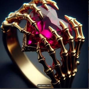Op de afbeelding: Een goudkleurige ring met een hartvormige, magenta edelsteen, vastgehouden door skeletachtige handen. De ring heeft een gedetailleerd ontwerp en een gepolijste afwerking. De tekst in de afbeelding luidt "Color: Golden / Size: 6".