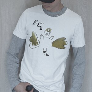 May include: A white t-shirt with a graphic of a cat with angel wings. The text "Angel Cat" is written above the cat. The person wearing the shirt is wearing a black baseball cap.