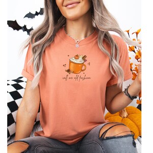 May include: A peach-colored t-shirt featuring a graphic of a mug of whipped cream-topped coffee with cinnamon sticks and a pumpkin. The text "call me old fashion" is below the mug.