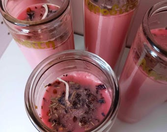 May include: Several pink candles in clear glass jars. The candles have a layer of dried herbs and flowers on top. The jars have gold lettering. The candles are arranged on a white surface.