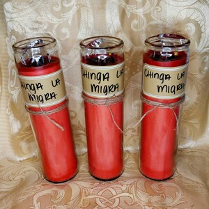 Op de afbeelding: Drie rode kaarsen in glazen potten met de tekst "Chinga La Migra" op een bruin label. Elke kaars is omwikkeld met touw. De kaarsen staan op een gouden, patroonachtige ondergrond.