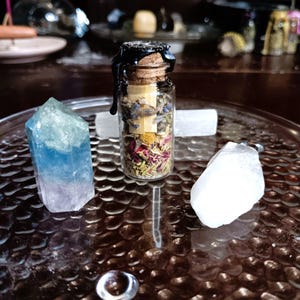 May include: A small glass bottle with a cork stopper sealed with black wax. The bottle is filled with dried herbs, flowers, and crystals. The bottle is sitting on a clear glass tray with two clear crystals.