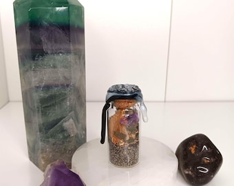 Puede incluir: Una pequeña botella de vidrio con un tapón decorativo, llena de hierbas y semillas secas, se encuentra sobre un soporte circular blanco. También hay un gran cristal verde y morado, una amatista morada y una piedra marrón oscura.