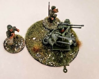 May include: A miniature wargaming scene featuring a green and brown anti-aircraft gun with two soldiers, one standing and one sitting, on a green and brown base.